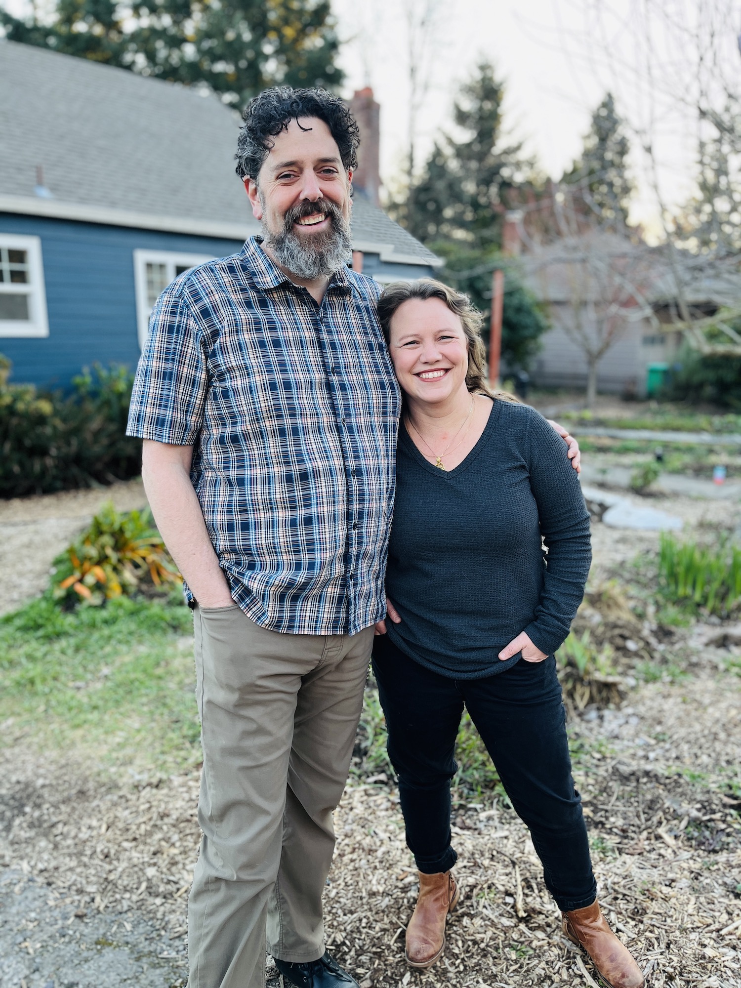 A tall, light-skinned man with a beard, and a shorter light-skinned woman smile and pose with their arms around each other in front of a blue house.