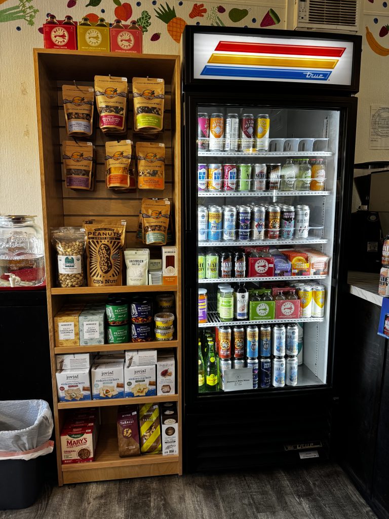 A refrigerated case and a display shelf in a neighborhood market are filled with locally-made foods and beverages for sale.