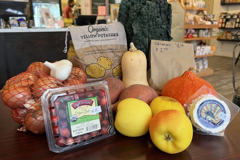 A collection of fall produce items arranged on a dark wood table: winter squash, apples, cranberries, kale, onions, and potatoes.