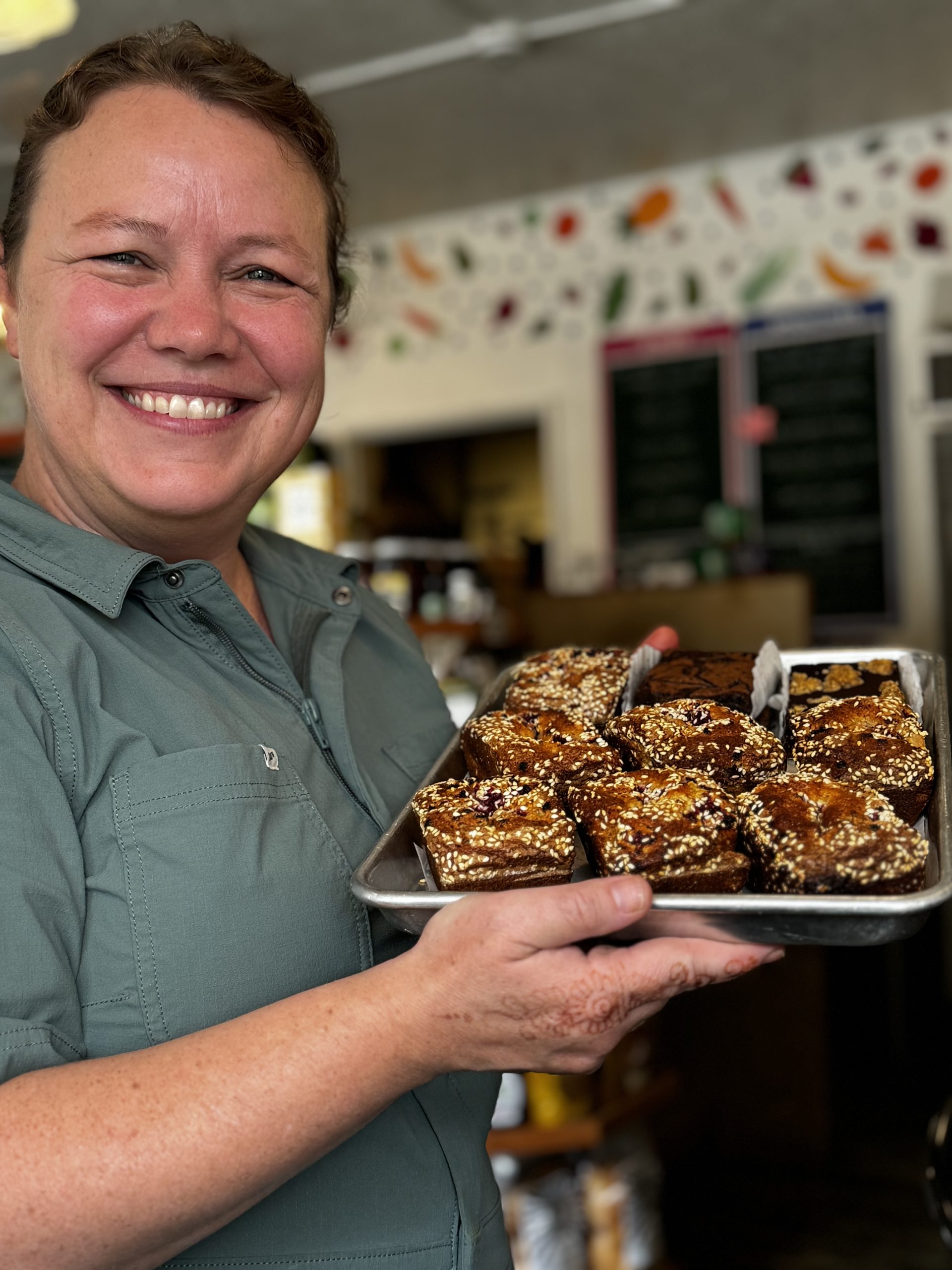 A smiling, light-skinned woman in a green coverall poses with a tray of pastries.