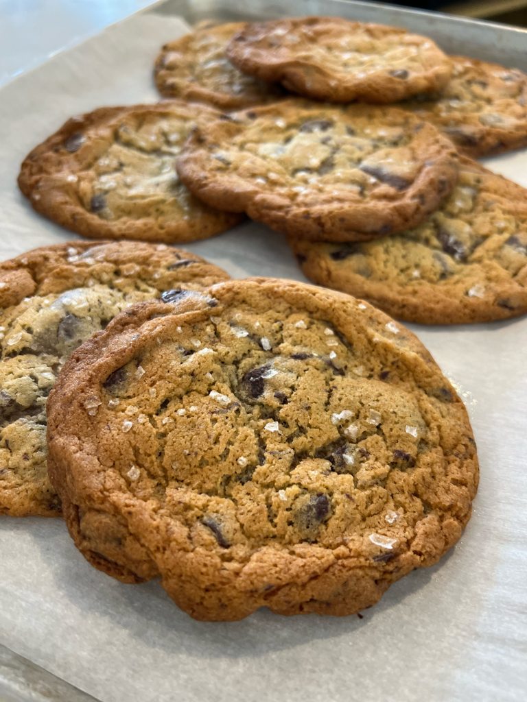 A paper-lined metal tray with fresh-baked chocolate chip cookies topped with flaky salt.