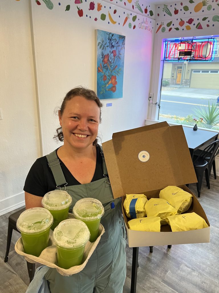 A smiling, light-skinned woman wearing overalls is holding a small catering order with a tray of green juices and a box with breakfast sandwiches wrapped in yellow paper.