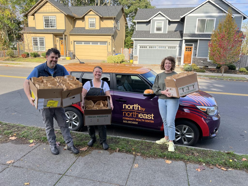 Three smiling light-skinned adults pose on the curb in front of a vehicle holding boxes full of bagged lunches. The car is from a community health center called North by Northeast.