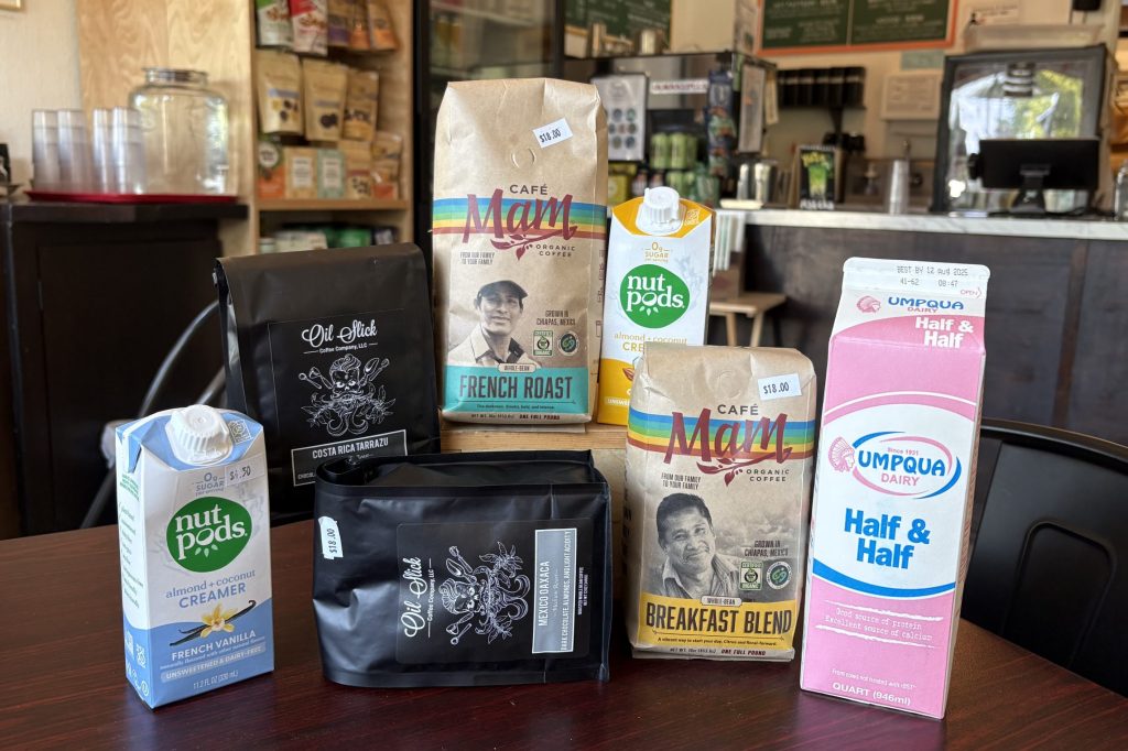 A collection of grocery items is arranged on a dark wood table: bags of small-batch coffee beans, flavored non-dairy creamers, and a carton of half-and-half.