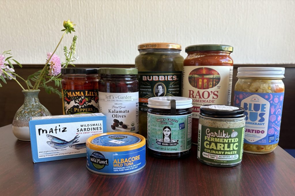 A collection of condiments and preserves is arranged on a dark wood table next to a vase of flowers: pickles, sauces, salsas, and tinned fish.