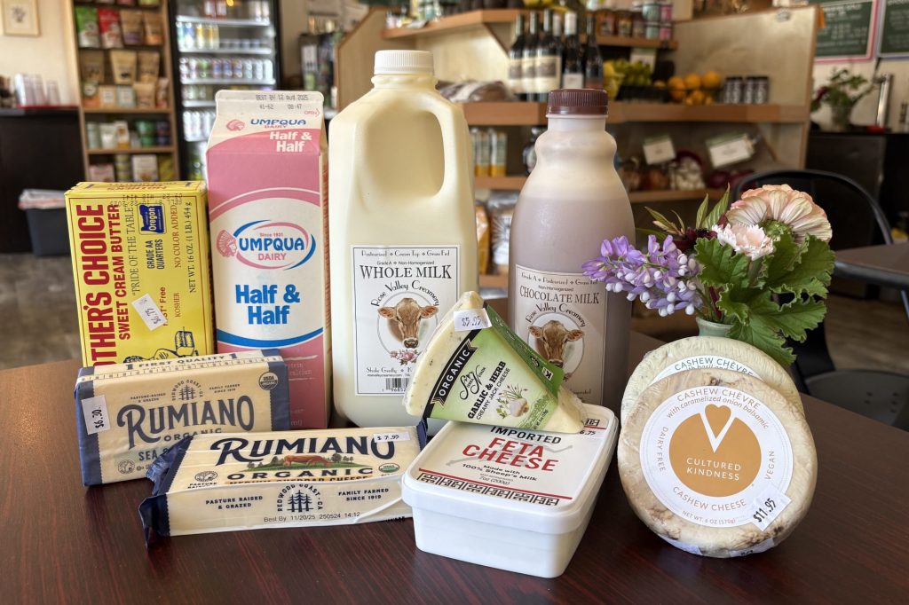 A collection of dairy products is arranged on a dark wood table next to a vase of flowers: milk, half and half, butter, and various dairy and non-dairy cheeses.