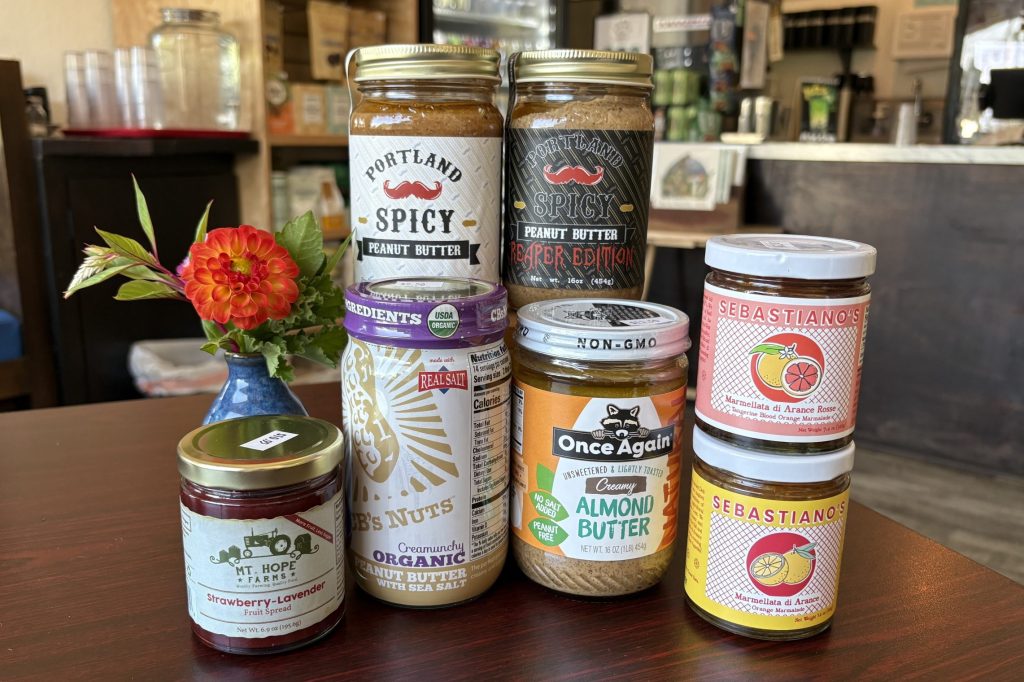 A collection of jars of nut butter and fruit jam are arranged on a dark wood table next to a vase of flowers.