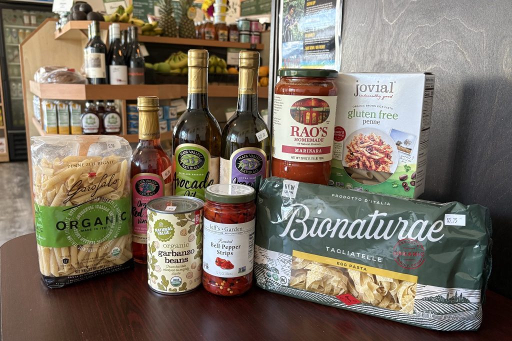 A collection of grocery items is arranged on a dark wood table: dried pasta, jars of sauce, bottles of olive oil and vinegar, and cans of beans and peppers.